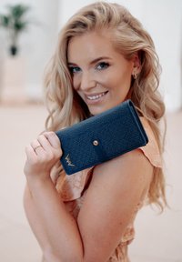 Dark blue, textured wallet with a gold logo detail and a snap closure, held by a person with wavy blonde hair.