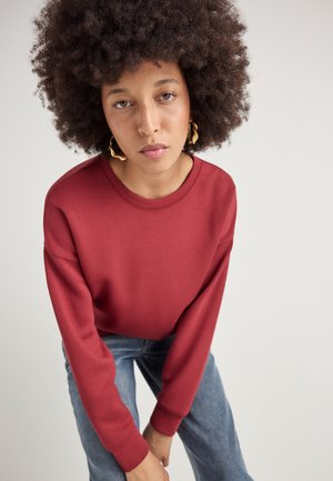 Red sweatshirt with long sleeves and a round neckline, paired with light blue jeans. The fabric appears smooth, featuring no visible patterns.