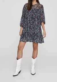 Šaty s květinovým potiskem z tmavé látky s nabíranými rukávy, délka po kolena, doplněné bílými kovbojskými botami s špičatými špičkami a dekorativním prošitím.