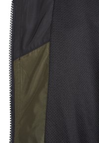 Gros plan d'une fermeture éclair noire à côté d'un tissu vert olive foncé et d'un matériau en maille noire texturée, montrant les détails du tissu et les coutures.