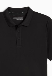 Zwart poloshirt met korte mouwen, twee knopen en een kraag, met Porsche Design en Puma branding aan de binnenkant van de hals.