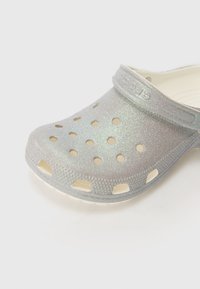 Zoccolo argento luccicante con fori di ventilazione, realizzato in materiale sintetico leggero, dotato di una fascia sul retro per una vestibilità sicura e suola texturizzata.
