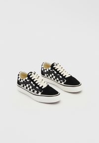 Paire de baskets basses avec un motif damier noir et blanc, des lacets blancs et des semelles épaisses blanches sur un fond uni.
