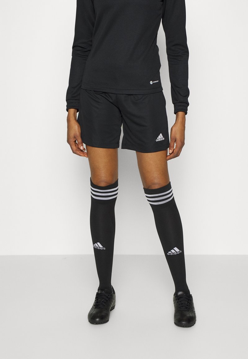 Persoon draagt een zwart shirt met lange mouwen, zwarte Adidas shorts, zwarte kniehoge sokken met witte strepen en zwarte sportschoenen, staand tegen een effen achtergrond.