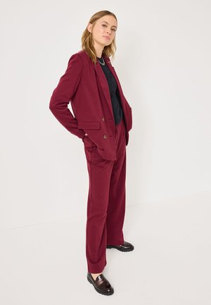 Costume ajusté bordeaux avec un blazer croisé et un pantalon large, associé à une chemise noire et des mocassins sombres brillants.