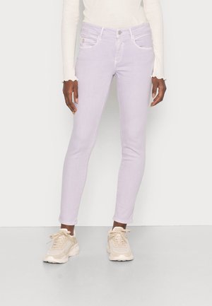 Person iført lyse lilla skinny jeans, hvid langærmet top med flæsekantede ærmer og beige chunky sneakers, stående mod en ensfarvet baggrund.