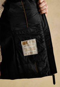 Dłoń trzyma otwartą czarną kurtkę, odsłaniając etykietę z tkaniny z marką Joules oraz tekstem o rzemiośle i wytrzymałości.