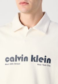 Polo majica boje kreme s mekim teksturom. Ima klasičan ovratnik i podebljani crni tekst: "calvin klein West 39th Street New York City."