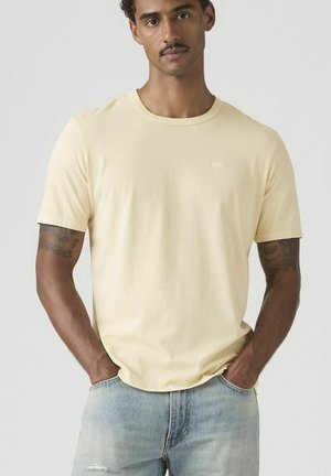 Hombre con tatuajes que lleva una camiseta lisa de manga corta color amarillo claro y jeans color azul claro, con las manos en los bolsillos, fondo neutral.