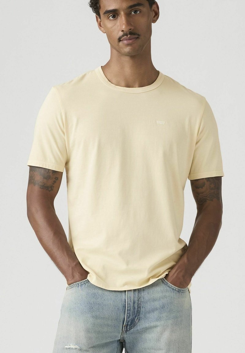 Uomo con tatuaggi che indossa una maglietta gialla chiara a maniche corte e jeans azzurri, mani in tasca, sfondo neutro.