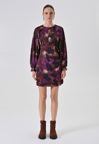 Kurzes Kleid aus dunkellila Stoff mit abstrakten Mustern in helllila und braun. Mit langen Ärmeln und gerafften Manschetten sowie einem runden Ausschnitt.