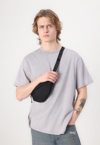 Svart crossbody bag med avrundet form og glidelås, med en teksturert rem merket "JACK & JONES." Båret over en lysegrå t-skjorte.