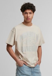 Beige bomull t-skjorte med avslappet passform, med en falmet grafisk trykk som leser "PROPERTY OF BASKETBALL" i blått. Korte ermer, rund hals.
