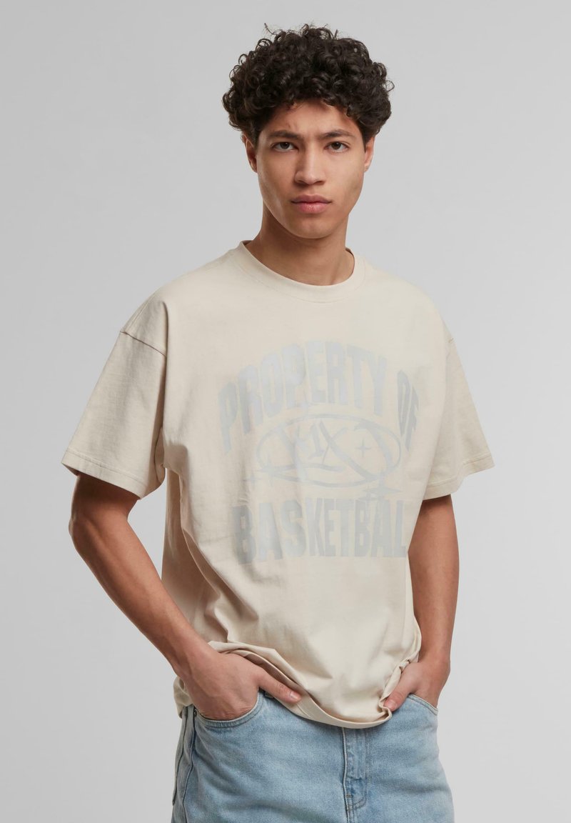 Beige bomull t-skjorte med avslappet passform, med en falmet grafisk trykk som leser "PROPERTY OF BASKETBALL" i blått. Korte ermer, rund hals.