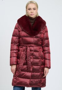 Vrouw in een glanzende bordeaux quilted jas met een grote nep-bont kraag en een tailleband, staand tegen een effen lichte achtergrond.