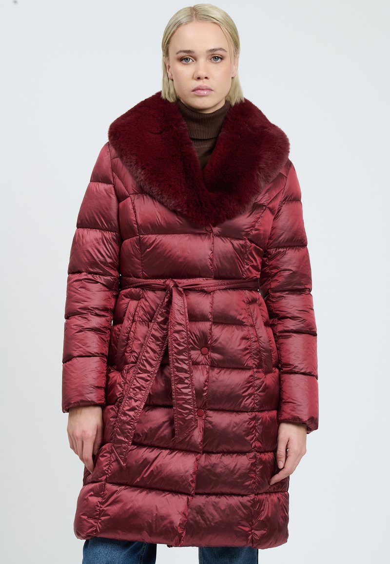 Vrouw in een glanzende bordeaux quilted jas met een grote nep-bont kraag en een tailleband, staand tegen een effen lichte achtergrond.