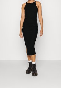 Femme portant une robe midi noire sans manches en maille côtelée avec une fente sur le côté, associée à des chaussettes blanches et des bottes noires à lacets épaisses.