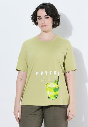 Kobieta z krótkimi ciemnymi włosami ma na sobie jasnozieloną koszulkę z grafiką matcha latte i napisem MATCHA LATTE.