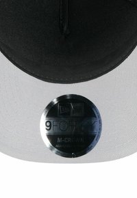 Schwarz-weiße Kappe mit einem verstellbaren Riemen, die eine strukturierte Krone und ein rundes Etikett mit der Aufschrift "9FORTY M-CROWN" auf der Innenseite aufweist.