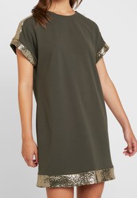 Sukienka w oliwkowym kolorze o luźnym kroju, z krótkimi rękawami i ozdobnym złotym wykończeniem rękawów oraz dołu. Wykonana z miękkiego, gładkiego materiału.