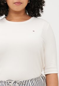 Person med krøllete hår som har på seg en lyse beige ribbet topp med en liten logo, kombinert med stripete bukser med snøring, plassert i sentrum av bildet.