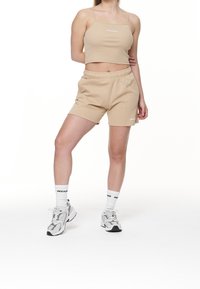 Kobieta wbeżowej bluzce typu crop top i szortach, w białych skarpetkach markowych oraz szaro-białych trampkach stoi na tle białym.