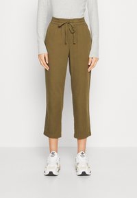 Pantalons courts amples de couleur vert olive, avec une taille élastique et un cordon de serrage, associés à des baskets blanches et beiges. Tissu à texture lisse.