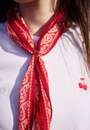 Écharpe rouge à motif paisley fixée par une bague autour du cou d'une personne, portée sur une chemise blanche avec une petite broderie de cerise rouge.