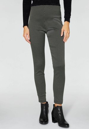 Beine in eng anliegenden schwarz-weißen Gemusterten Hosen, kombiniert mit schwarzen Stiefeletten mit Absatz, stehend vor einem schlichten hellen Hintergrund.