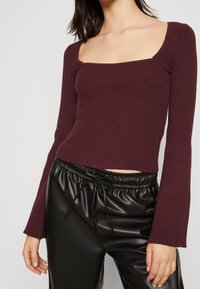 Bordowa prążkowana bluzka z długim rękawem o kwadratowym dekolcie, dopasowanym kroju i teksturowanej tkaninie, zestawiona z czarnymi spodniami z elastycznym pasem.
