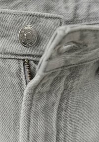 Gros plan sur un tissu en denim gris clair montrant un bouton en métal étiqueté "Authentic Denim MNG" et une fermeture éclair partiellement ouverte.