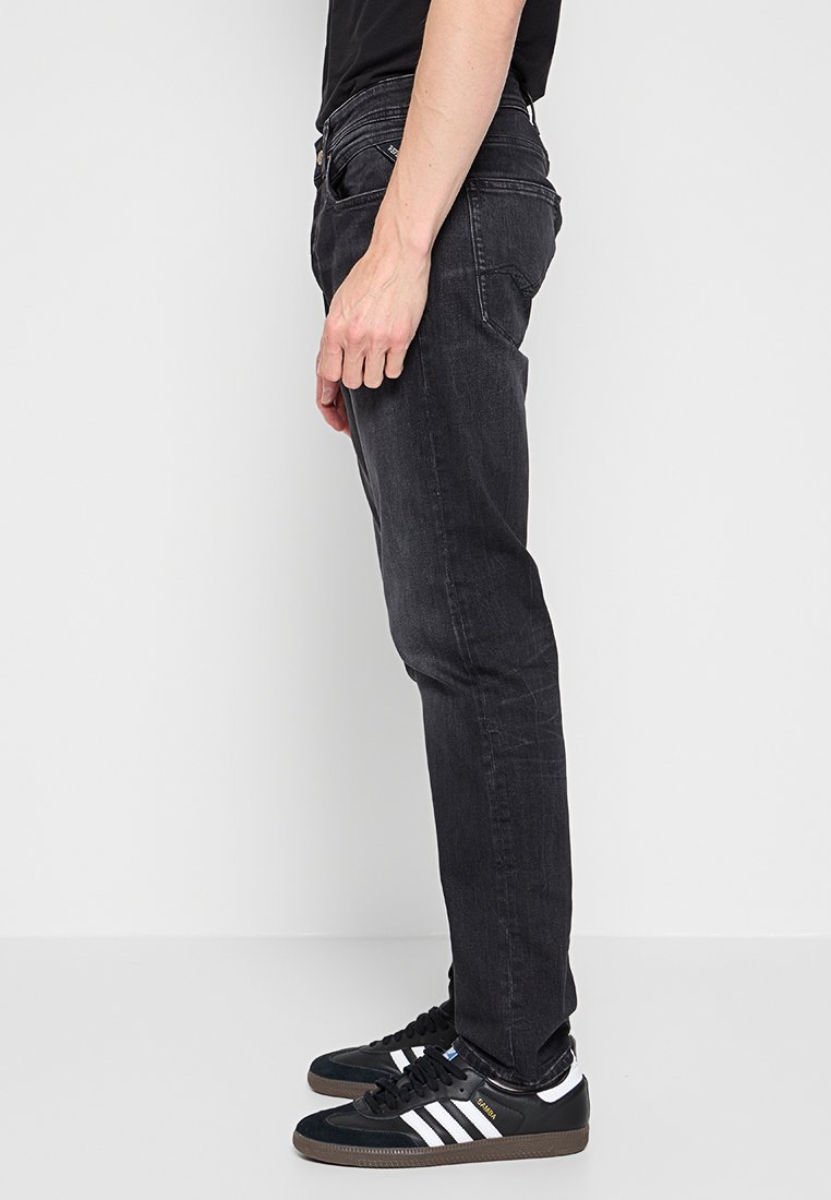 Persoon in zwarte slim-fit jeans en zwarte Adidas sneakers met witte strepen, staand tegen een effen witte achtergrond.