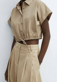 Camicia beige a maniche corte con bottoni, con orlo elastico, abbinata a pantaloni plissettati beige a vita alta.
