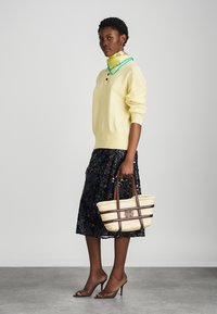 Geel gebreid sweater met een hoge kraag en knopen detail, gecombineerd met een donkere bloemenrok. Houdt een strohandtas vast met bruine accenten.