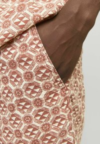 Tissu à motifs présentant des dessins géométriques et floraux dans des couleurs rouille et crème. Le matériau semble léger et texturé, avec une poche visible.