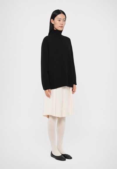 Pull en tricot noir à col roulé, coupe ample, associé à une jupe beige clair mi-genou, des collants blancs et des ballerines noires. Design simple et minimaliste.
