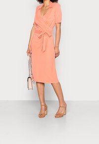 Žena stojí v korálových šatech s páskem v pase, hnědých sandálech na blokovém podpatku a drží světle růžovou kabelku s řetězovými popruhy.