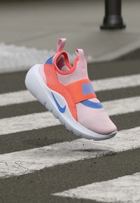 Nike-Sneaker mit einem rosa Mesh-Obermaterial, orangenem Riemen mit Logo, blauen Akzenten und einer klobigen weißen Sohle, vor einem Zebrastreifen-Hintergrund.