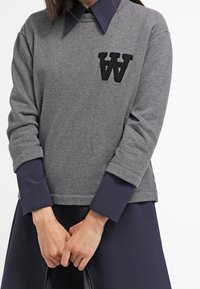 Harmaa huppari tummansinisellä kauluksella, jossa on brodeerattu musta "W" vasemmassa rinnassa. Pitkät hihat tummansinisillä resoreilla. Sujuva pinta.