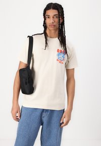 Beige cotton t-shirt with graphic print in red and blue, paired with blue denim shorts. Black crossbody bag with zip closure worn over the shoulder.
