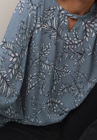 Blusa azzurro chiaro con motivo floreale in blu scuro e bianco, caratterizzata da un collo a chiave e maniche lunghe e ampie. Tessuto strutturato.
