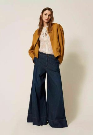 Donna con giacca color senape sopra una camicetta bianca in pizzo, abbinata a jeans a vita alta a gamba larga di colore blu scuro, in piedi davanti a uno sfondo semplice.