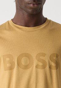 Mustard yellow cotton t-shirt with a crew neck; features large, textured "BOSS" logo in a slightly darker shade. Smooth fabric texture.