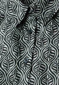 Zwart-wit stof met een bladpatroon en een zachte, gestructureerde textuur. Heeft een kraag en drie donkere knopen aan de voorkant.