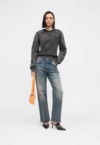 Μαύρο φούτερ με υφή στο ύφασμα και ραφές σε αντίθεση, συνδυασμένο με ανοιχτόχρωμα τζιν παντελόνια. Το μοντέλο κρατά μια μικρή πορτοκαλί τσάντα.