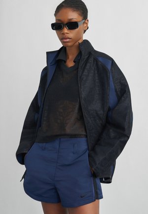 Femme portant des lunettes de soleil noires, un haut noir transparent, une veste zippée noire et marine, et un short marine avec le logo Nike, debout les mains dans les poches.