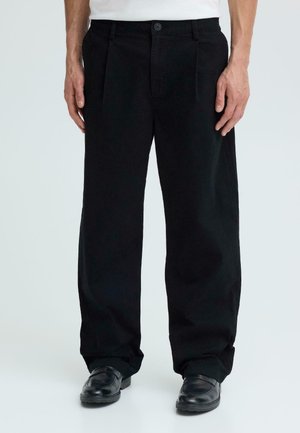 Mężczyzna ubrany w czarne, luźne spodnie i czarne skórzane buty, stojący na tle jednolitego jasnego tła.