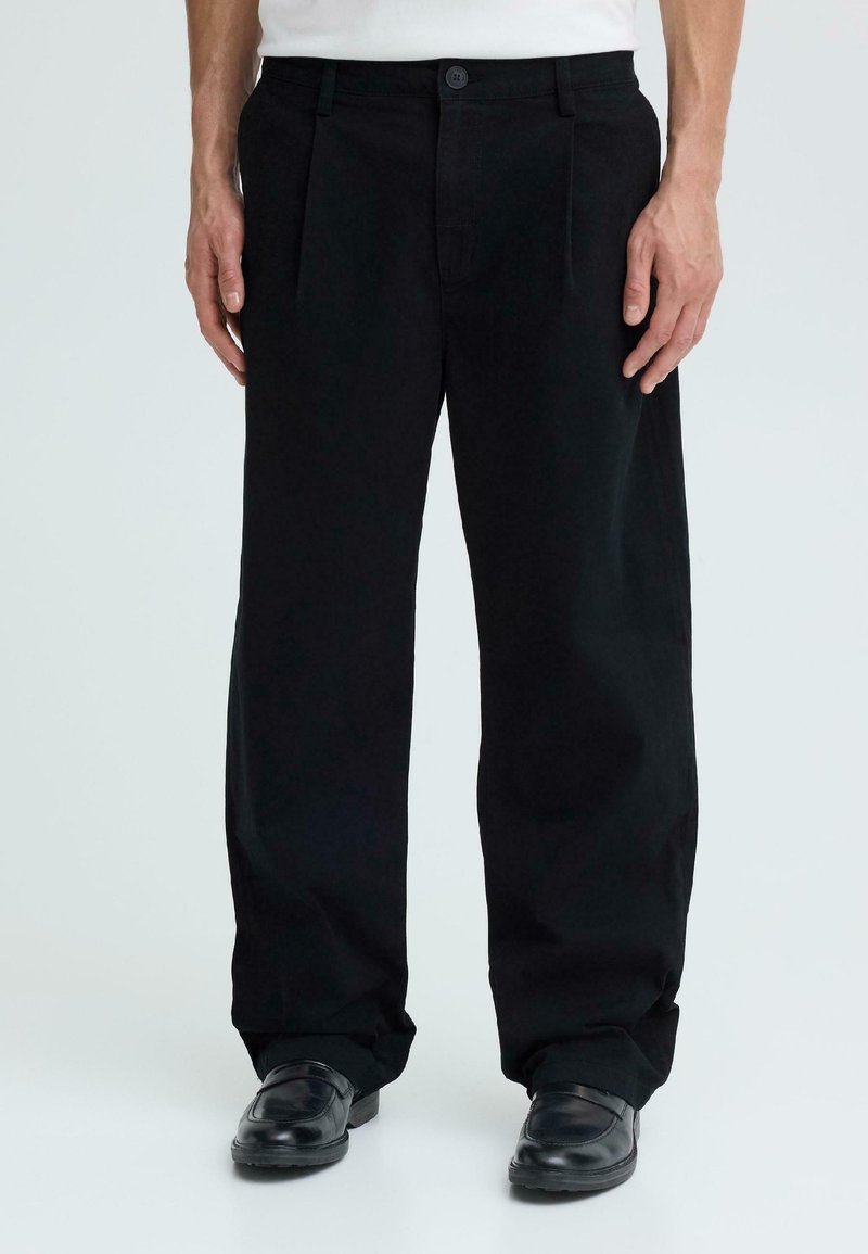 Homme portant un pantalon ample noir et des chaussures en cuir noires, debout devant un fond clair uni.