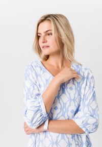 Hellblau und weiß gemusterte Bluse mit V-Ausschnitt, gerafften Ärmeln und weichem Stoff. Das Design weist durchgängig geometrische Formen auf.