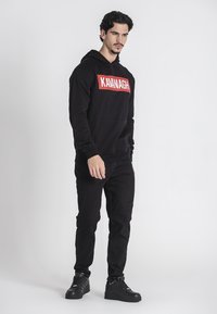 Čierna mikina s kapucňou s nápisom "KAVANAGH" v tučnom červenom písmene na prednej strane, skombinovaná s čiernymi džínsy a čiernymi teniskami. Kapucňa je nastaviteľná.
